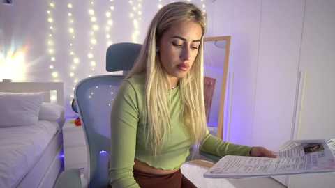 Media: Video of a young blonde woman in a green top, reading a magazine in a modern bedroom with white walls, fairy lights, and a bed in the background.