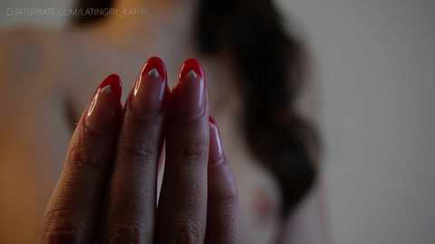 Media: A close-up video of a woman's hand with neatly manicured nails painted in a glossy red polish. The background is blurred, featuring a woman with long, dark hair and visible cleavage, suggesting a suggestive, intimate setting.