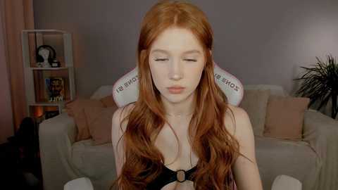 Media: Video of a young, fair-skinned woman with long, wavy red hair, wearing a black bra, sitting in a gaming chair in a cozy living room.