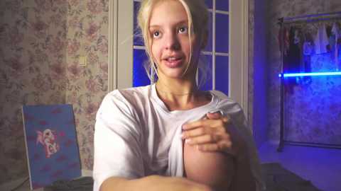 Media: Video of a young, blonde, fair-skinned woman with large breasts, partially lifting her white t-shirt to expose one breast. She's indoors, in a dimly lit room with floral wallpaper and a blue-framed painting.