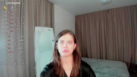 Media: Video of a young woman with fair skin, long brown hair, and a V-shaped face, wearing a black jacket, in a beige room with a bed and curtains.