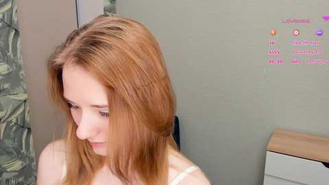 Media: Video of a fair-skinned, young woman with long, straight, reddish-brown hair styled in a side part. She wears a white tank top, standing indoors with a light green wall and a camo-patterned curtain in the background.