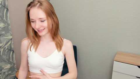 Media: A video of a slender, fair-skinned young woman with long, straight auburn hair, wearing a white crop top, seated indoors against a muted green wall, smiling.