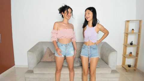 Media: Video of two smiling young women in pastel outfits, standing in a modern living room with a beige couch, wooden shelf, and a brown wall.