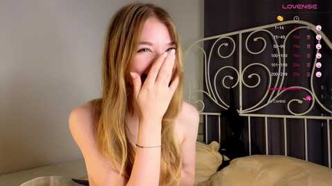 Media: Video of a topless, fair-skinned, blonde woman with long hair, covering her face with her hands, in a bedroom with a white wrought-iron bed and black wall with a pink calendar.