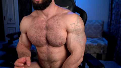 Media: Video of a shirtless, muscular man with a beard, sitting on a gaming chair in a dimly lit room. He has a tattoo on his right shoulder and a hairy chest.