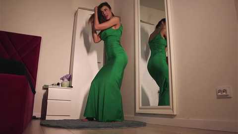 Video of a woman in a green, floor-length dress, standing in a dimly lit bedroom, adjusting her hair in front of a full-length mirror, with a bed and dresser visible.