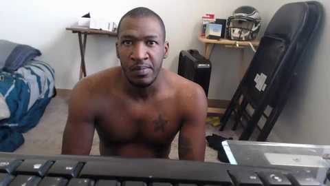 Media: Video of a shirtless, muscular Black man with a bald head, sitting on the floor, looking directly at the camera. He is in a cluttered room with a football helmet, a blue bag, and a black keyboard in the foreground.