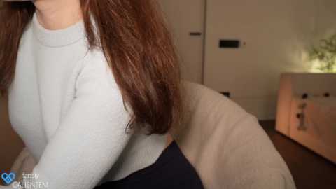 Media: Video of a woman with long, wavy brown hair wearing a light gray, long-sleeved sweater. Background shows a modern, minimalist room with a white door, a white cabinet, and a potted plant.