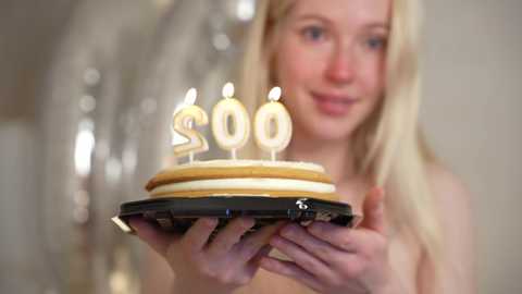 Media: Video of a blonde woman holding a cake with \"30\" candles, smiling in a festive indoor setting with silver balloons and soft, blurred background.