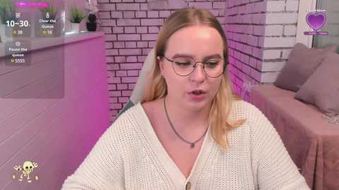 Media: Video of a blonde woman with glasses, wearing a cream cardigan, sitting on a couch, in a modern, purple-themed room with brick walls and potted plants.