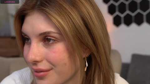 Media: Video of a smiling young woman with fair skin, straight brown hair, and subtle makeup, wearing a white top, in a modern living room with hexagonal wall art.