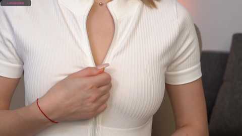 Media: Video of a woman with fair skin, wearing a white, ribbed, short-sleeved top, unzipping it with her left hand, revealing a red bracelet.