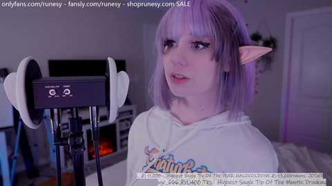 Media: Video of a young woman with pale skin, purple hair, and pointy elf ears, wearing a white hoodie with a colorful graphic, seated in front of a microphone in a dimly lit room.