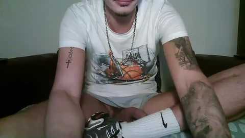 Media: Video of a tattooed, light-skinned man with a trimmed beard, wearing a white basketball T-shirt and white socks, sitting on a dark couch, displaying an erect penis.