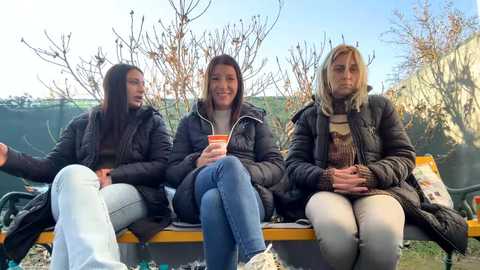 Media: Video of three women sitting on a wooden bench in a garden, wearing winter coats and scarves. They hold red mugs, smiling, with bare trees and a green background.