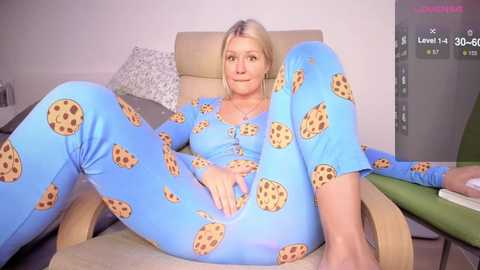 Media: Video of a blonde Caucasian woman in blue pajamas adorned with chocolate chip cookies, reclining on a beige chair in a modern living room.