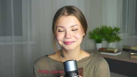 Media: Video of a young woman with fair skin, brown hair, and a nose ring, smiling while recording herself with a microphone. Background features a green plant and white curtains.