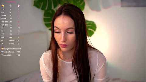 Media: A video of a young Caucasian woman with long brown hair, wearing a sheer white top, sitting indoors, with a green plant in the background.