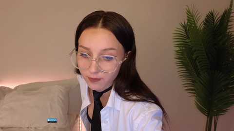 Media: Video of a young Asian woman with long dark hair, wearing glasses and a white shirt, smiling, with a green plant in the background.