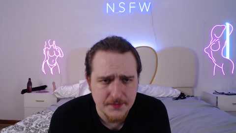 Media: Video of a bearded man in a black shirt, sitting in a bedroom with neon nude figures on the wall, messy bed, and a bottle.