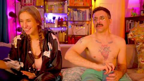 Media: Video of a laughing woman with braids, wearing a black robe, and a mustachioed man with tattoos, both sitting on a couch in a colorful, cluttered room.