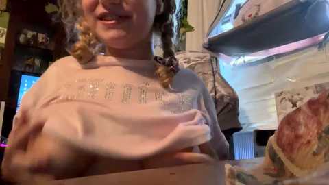 Media: Video of a young girl with blonde braided pigtails, wearing a light pink shirt, holding a partially eaten ice cream cone in a cozy, dimly lit kitchen with a refrigerator and cabinets.