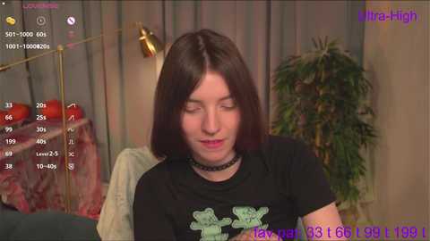 Media: Video of a fair-skinned woman with straight, shoulder-length brown hair, wearing a black graphic tee with green bear illustration, black choker, and a subtle smile. Background includes a potted plant, wall-mounted lamp, and a red apple on a table.