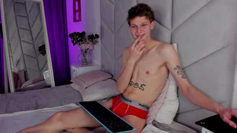 Media: Video of a young, slim, shirtless man with short brown hair, wearing red Calvin Klein underwear, sitting on a bed with a computer keyboard, in a modern, purple-lit bedroom.