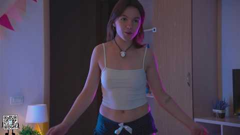 Media: Video of a slender, young Asian woman with light skin and straight brown hair in a white tank top and blue shorts, standing in a dimly-lit bedroom.