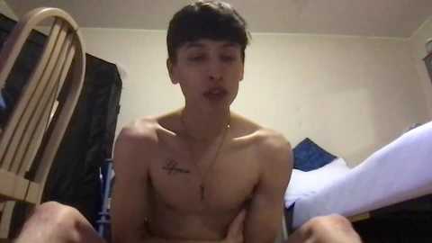 Media: Video of a shirtless teenage boy with a thin build, short dark hair, and a tattoo on his chest, sitting on a bed in a dimly lit room.