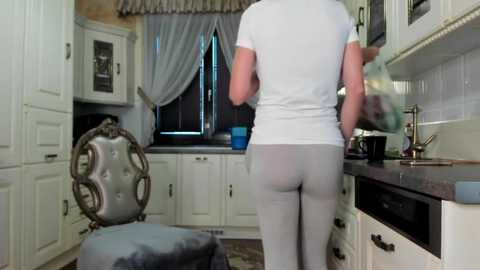 Media: A video of a person in a white t-shirt and grey pants standing in a kitchen, facing away, with a vintage chair and white cabinets in the background.