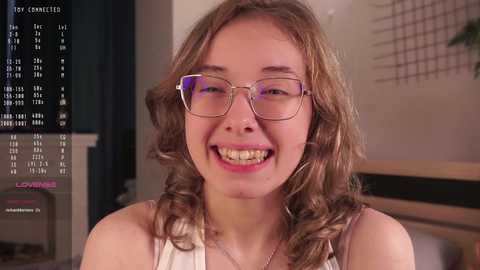 Media: Video of a smiling, young woman with braces, wearing glasses and a white sleeveless top. Background shows a TV screen displaying a calendar and a wall with a grid pattern.