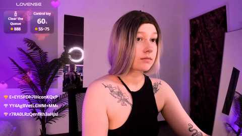 Media: Video of a young woman with shoulder-length, light brown hair, wearing a black tank top, showing tattoos on her shoulders. She's indoors with purple lighting, surrounded by plants and wall art.