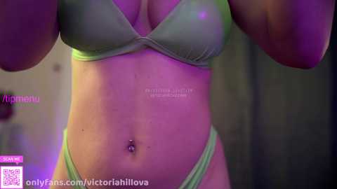 Media: Video of a light-skinned woman in a green bra and matching thong, with a navel piercing, taken indoors with purple lighting.