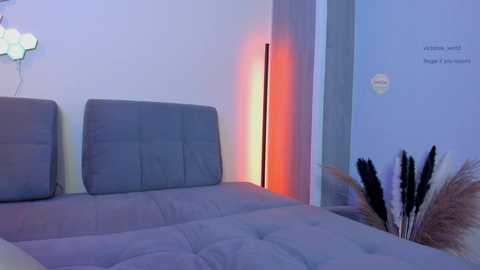 Media: Video of a minimalist bedroom with a gray upholstered bed, a modern floor lamp with a red light, and a vase of dried pampas grass on the right. The walls are light blue, and the room has a serene, contemporary ambiance.