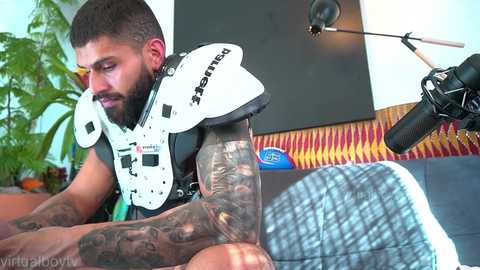 Media: Video of a bearded, tattooed man with a muscular build wearing a white, black, and red helmet, sitting in a room with a green plant, a striped rug, and a black microphone stand.