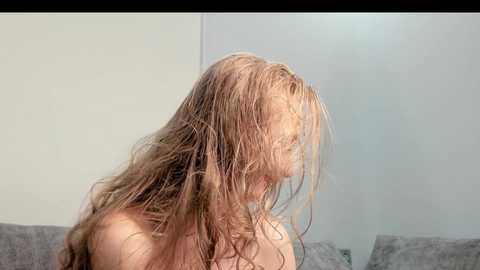 Media: A video of a young, fair-skinned woman with long, wet, light brown hair covering her face, standing topless against a light-colored wall.