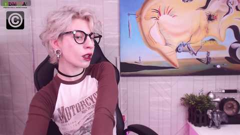 Media: Video of a pale, short-haired woman with glasses, black choker, and brown tee, sitting in a chair with a colorful, surreal painting behind her.