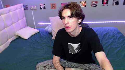 Media: Video of a young man with fair skin, brown hair, and a neutral expression, sitting on a blue bedspread in a bedroom with white furniture and a wall of framed photos.