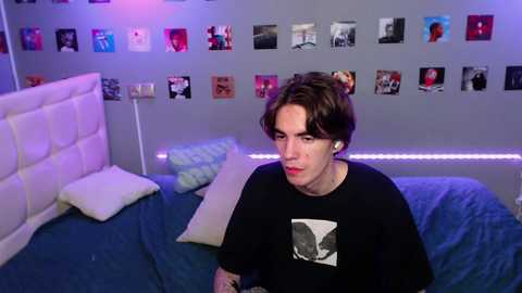 Media: Video of a young man with short brown hair, wearing a black graphic t-shirt, sitting on a bed in a brightly lit, modern bedroom with a gray wall adorned with a collage of colorful photos.