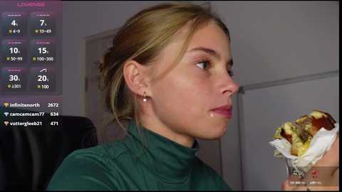 Media: Video of a young woman with light skin and blonde hair in a bun, wearing a green turtleneck, eating a burger. Background includes a pink chat window with social media notifications.