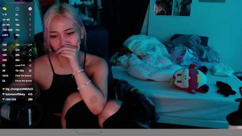 Media: Video of a young, blonde woman with a thin build, wearing a black tank top, sitting on a bed with a plush toy and messy blankets. Dimly lit room, blue screen overlay with social media notifications.