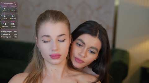 Media: Video of two women, one blonde with light skin, one brunette with medium skin tone, hugging closely, both topless, in a dimly lit room with a green couch in the background.