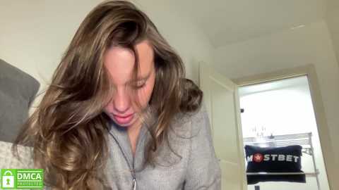 Media: A video of a young woman with long, wavy brown hair, wearing a light grey zip-up hoodie, looking down with a slight smile. The background shows a white door and a grey pillow.