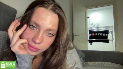 Media: Video of a young woman with long brown hair, wearing a gray sweater, lying on a gray bed, looking tired and resting her head on her hand. Behind her, a door is open, displaying a hanging black \"STREET\" t-shirt.