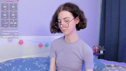 Media: Video of a young, pale-skinned, short-haired woman with glasses, wearing a light grey shirt, standing in a purple-lit bedroom with a blue bedspread and a digital temperature display on the wall.