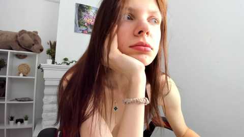 Media: Video of a young Caucasian woman with long brown hair, light skin, and green eyes, resting her chin on her hand. She wears a pink top and a white scrunchie on her wrist. Background features a white wall, a stuffed bear, and a colorful painting.