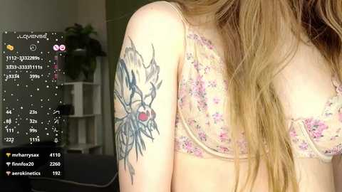 Media: A video of a young woman with long, wavy blonde hair, wearing a floral lace bralette. Her left arm displays a detailed tattoo of a dragon. The background shows a potted plant and a white shelf.