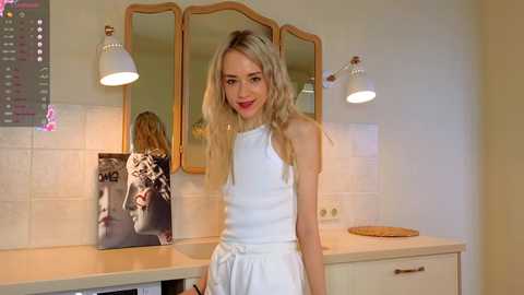 Media: Video of a blonde woman in a white dress standing in a modern kitchen with beige tiles, a countertop, and two pendant lights.